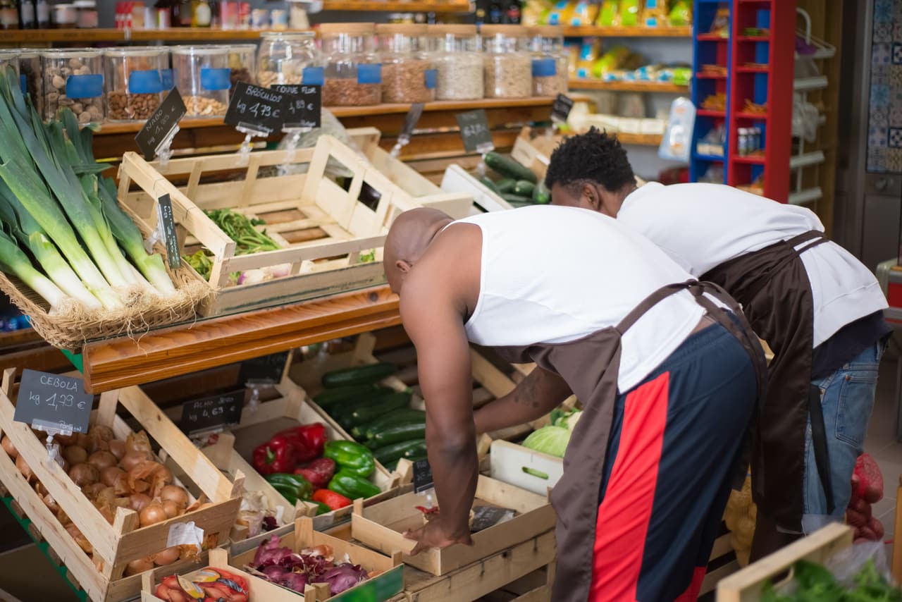 Dois comerciantes arrumando verduras e legumes nos caixotes de madeira de um hortifrúti
